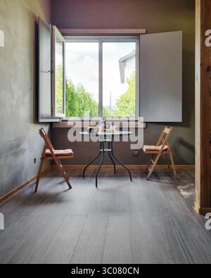 Cozy dining nook with concrete walls, open window, round glass table, two wooden folding chairs, dark hardwood floors. Stock Photo