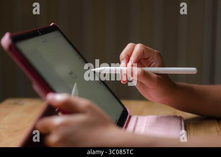 Cute Asian girl drawing using electronic tablet and stylus pen. Asian elementary school girl using digital tablet sitting in living room at home. Stock Photo