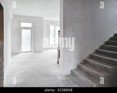 Interior of a new house under construction. Interior of unfinished house: empty room with large windows and stairs leading to second floor. Stock Photo