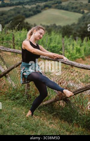 Woman gracefully practicing ballet exercises outdoors using a wooden fence as a support. Her attire is colorful and suitable for dance, set against a Stock Photo