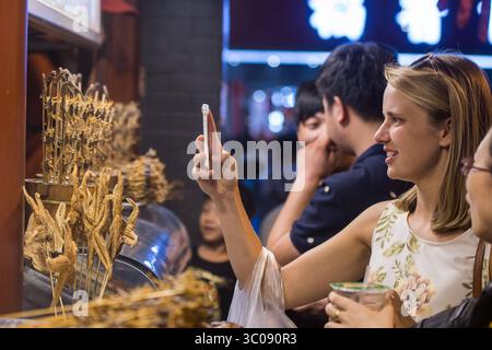 June 25, 2016 - Beijing, China - Beijing, China - Female tourist taking a picture on her cell phone of scorpions and starfish on sticks for sale at the Donghuamen Snack Night Market, a large outdoor market that is an attraction for locals and tourists, located at the north entrance of Wangfujing Street in Beijing. (Credit Image: © Edwin Remsberg / Vwpics/VW Pics via ZUMA Wire) Stock Photo
