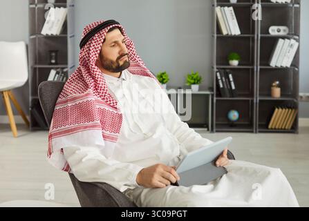 Thoughtful Arabian man posing in armchair with digital tablet, surfing internet or working online, Stock Photo