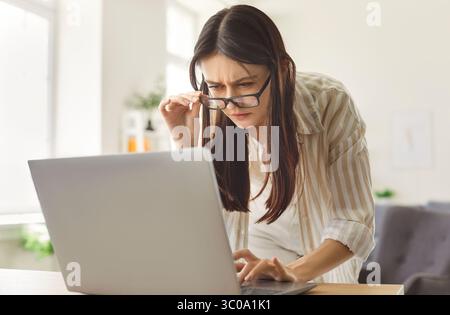 Concentrated woman focused in screen of laptop, reading something scrupulously, wearing spectacles Stock Photo