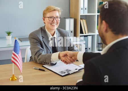 Shaking hands across desk, people sitting greeting in office near American flag, woman smiling Stock Photo