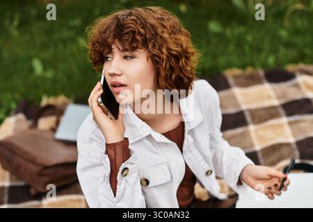 A young woman with curly hair sits on a blanket in the park, enjoying the summer sun. Stock Photo