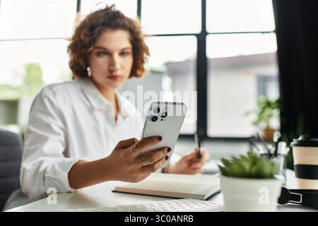 Curly-haired businesswoman focuses on her smartphone while working in a stylish office. Stock Photo