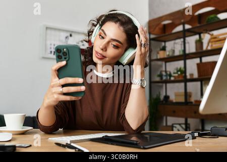 Focused young woman with curly hair edits photos at her home workspace while wearing headphones. Stock Photo