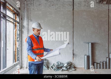 A construction worker wearing a safety vest and helmet focuses on building plans inside a partially finished office. The space features exposed brick Stock Photo
