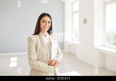 Smiling female realtor in bright empty real estate holding clipboard, looking cheerfully at camera. Stock Photo