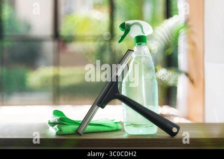 Window cleaner spatula, spray product and microfibre cloth on wooden table and background window. Front view. Stock Photo