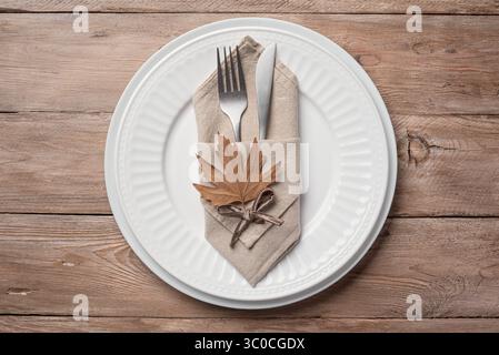 Autumn elegant place setting with dry maple leaves on wooden background, top view. White autumn minimal Thanksgiving table setting template. Stock Photo