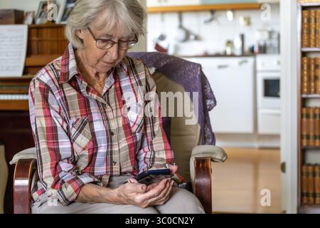 An elderly, gray-haired woman sits thoughtfully looking down at the screen of her mobile phone. Stock Photo