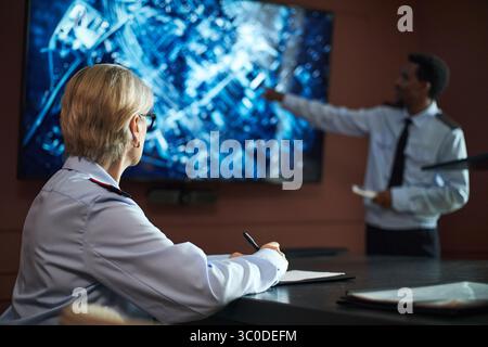 Middle aged Caucasian woman sitting at desk writing notes, while middle aged Black man standing in background pointing at large digital screen displaying city map during meeting Stock Photo