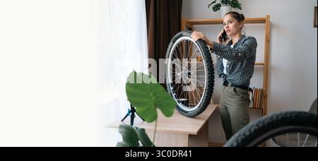 woman repairing bicycle at home by herself holding wheel and speaking on phone asking for help and consultation, concept of self-repair, communication Stock Photo