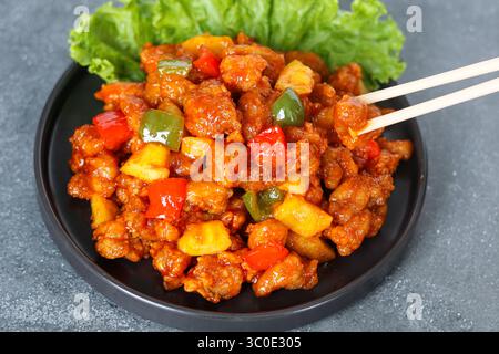 Sweet and sour chicken served on a dark plate, with a pair of chopsticks picking up a piece. Decorated with bell peppers and pineapple chunks. Stock Photo