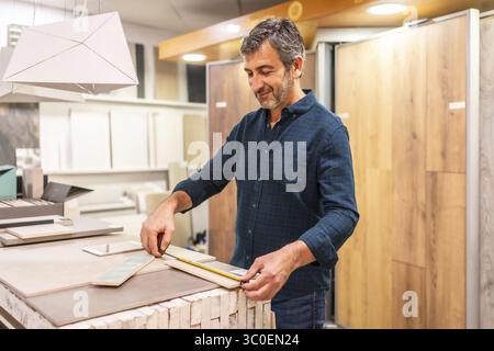 Smiling architect taking measurements of ceramic tile samples using a cutter in a home improvement store, during renovation or construction project Stock Photo