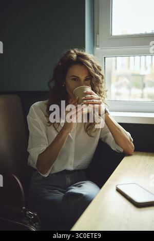 A woman with curly hair sits by a window in a cafe, holding a warm cup of coffee. Sunlight streams in as she enjoys her drink, dressed casually in a w Stock Photo