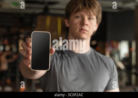 Athlete standing and displaying cellphone with blank screen in blurred gym setting. Fitness center. Mock up. Space for design. Stock Photo