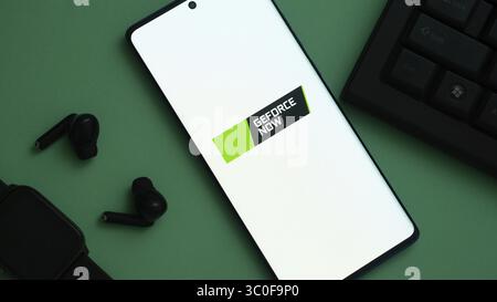 Smartphone with 'GEFORCE NOW' logo on green surface, surrounded by earbuds, watch, and keyboard Stock Photo