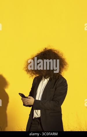 Vertical portrait of unrecognizable long-haired businessman with cellphone over yellow background Stock Photo