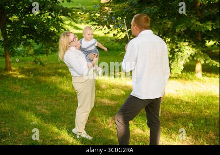 Happy family is relaxing in park. Father is happily playing and blowing soap bubbles, mother is holding cute baby in arms, laughing with him, catching Stock Photo