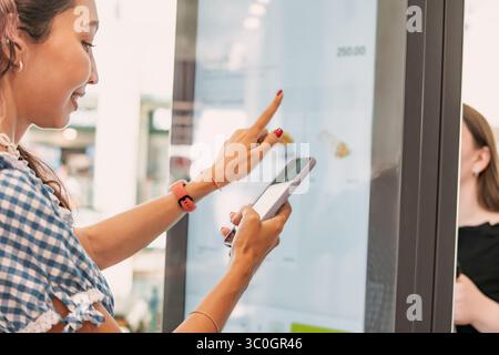 Customer using smartphone and interactive self ordering kiosk at fast food restaurant to choose meal and make contactless payment Stock Photo