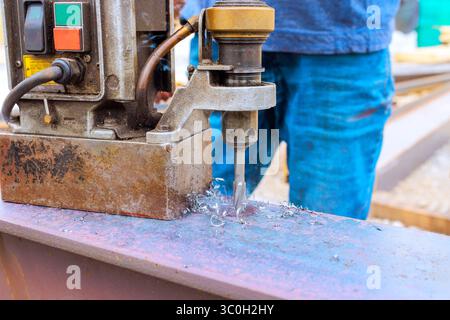 Worker operates metal drilling machine on steel beams while shavings fly everywhere in an industrial area. Stock Photo
