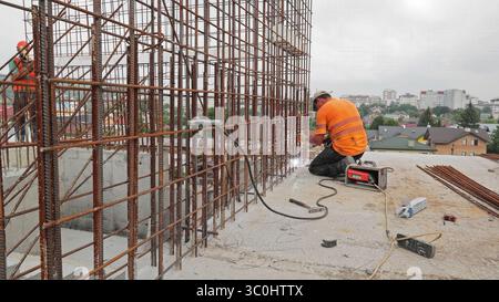 Construction Worker Welding Steel Reinforcement Framework. A construction worker wearing an orange safety shirt and helmet welds a large steel Stock Photo