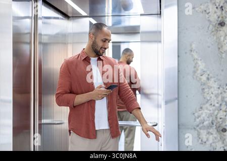 A man stands in an elevator, using his phone and pressing a button, with a reflective mirror on the side. Stock Photo