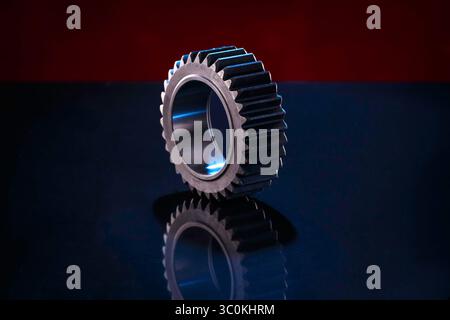 A close-up studio shot of a large, metallic gear resting on a dark, reflective surface. The gear is predominantly dark gray with a polished finish, ex Stock Photo