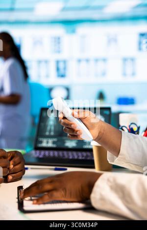 Female doctor measuring the temperature during medical consultation at the cabinet, using a digital thermometer tool to check for flu symptoms. Health expert ensures diagnosis for a patient. Stock Photo