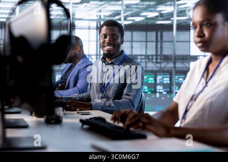 Cheerful admin next to colleagues doing yearly checkup to prolong data center electronics life span and avoid downtime. Happy engineers in server room monitoring server infrastructure components Stock Photo