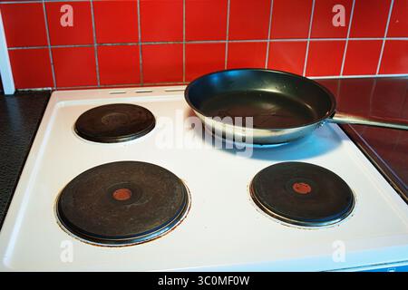 Black nonstick frying pan placed on a white electric stove with four round hotplates in a domestic kitchen with the vibant red tiled backsplash Stock Photo
