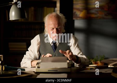 Senior Caucasian man sitting at desk using typewriter, concentrating on typing document, bookshelf and painting in background, wearing formal shirt and vest, working alone indoors Stock Photo