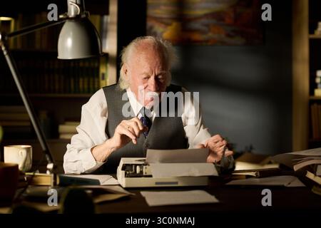 Senior Caucasian man sitting at desk using typewriter, holding paper and appearing focused, surrounded by documents and bookshelves, working in office or study environment Stock Photo