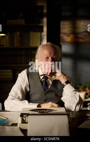Portrait of senior Caucasian man sitting at desk holding eyeglasses, resting chin on hand, looking into camera, surrounded by papers and vintage typewriter in library setting Stock Photo