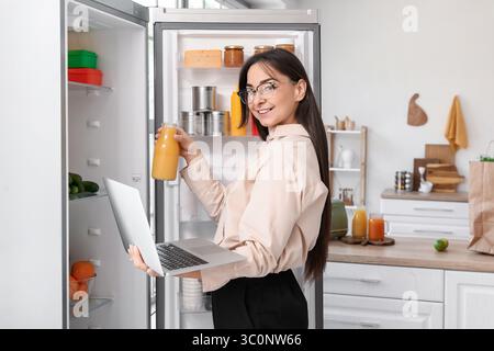 Beautiful businesswoman with laptop taking juice from open fridge in kitchen Stock Photo