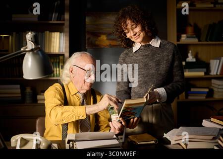 Senior Caucasian man sitting at desk reading book with young adult Caucasian woman standing beside him smiling, both engaging in discussion surrounded by bookshelves and papers Stock Photo