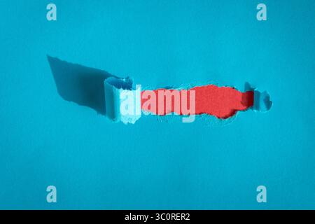 Hole ripped in blue paper on red background. Copy space. Stock Photo