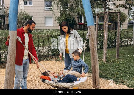 Happy family with two sons playing together on a large rope swing at playground. Parents smiling, bonding and spending quality time with kids outdoors Stock Photo