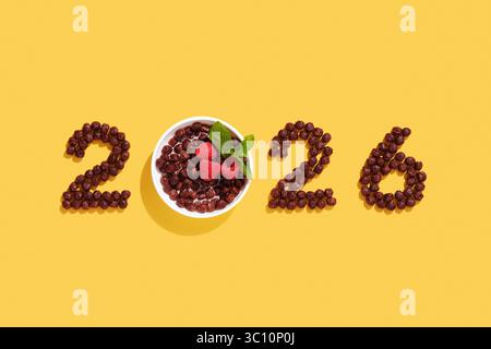 Cereal numbers form 2026 with chocolate balls and raspberry in a bowl. New year breakfast concept on yellow background with food design elements. Stock Photo