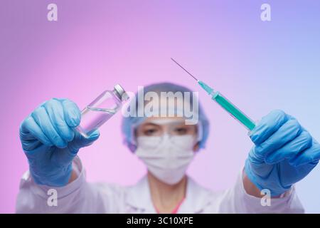 Modern Medical worker in white coat and protective gear holding vaccine vial in one hand and syringe in other hand on neon background, modern medicine Stock Photo