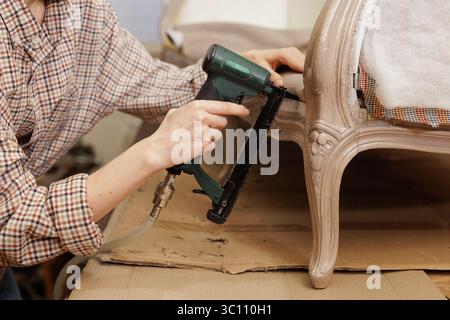 Making new upholstery on old restored furniture. A woman works with a pneumatic stapler in her workshop. Furniture repair Stock Photo