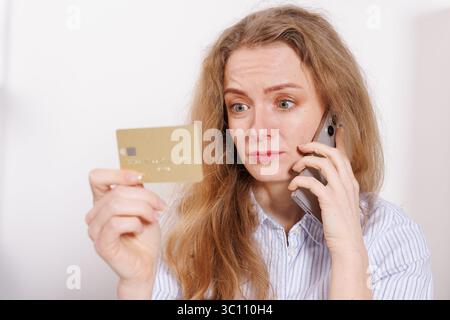 Confused woman with credit card talking on phone at home. Be careful - fraud Stock Photo