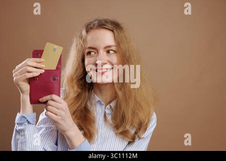 Smiling woman takes out gold bank card from red wallet on uniform beige background, online payments, virtual money, keeping money in bank Stock Photo