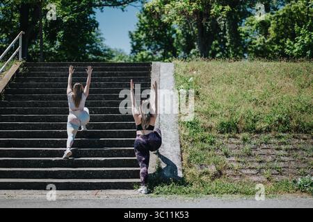 Two women performing yoga exercises outdoors on stairs surrounded by greenery Stock Photo