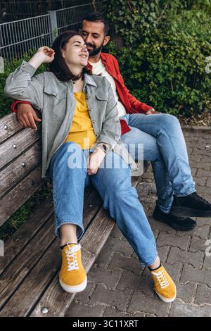 Interracial couple sits together on a park bench, smiling and enjoying a peaceful moment. A warm, loving connection capturing diversity, unity, and ha Stock Photo