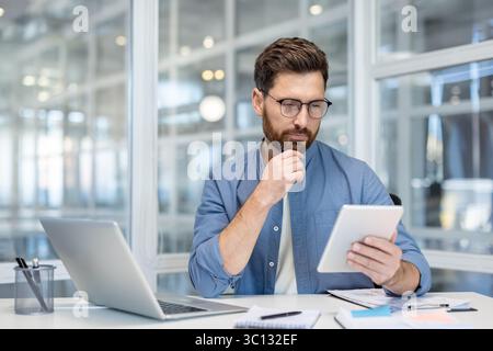 A thoughtful businessman in glasses examines his tablet in an office setting, with a laptop and documents nearby. Stock Photo