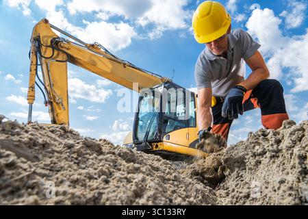 A construction worker is kneeling in the sand, using his hands to dig while an excavator operates nearby under clear skies. Stock Photo