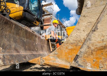 A construction worker in a helmet takes a break while seated on heavy machinery at a busy building site under a clear blue sky with clouds. Stock Photo
